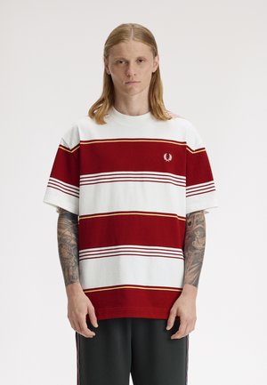 Jeune personne aux cheveux longs, portant un t-shirt rayé rouge et blanc et un pantalon noir, se tenant devant un fond uni.