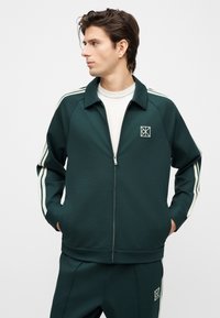 Νεαρός άνδρας που φοράει σκούρο πράσινο φόρμα με φερμουάρ και λευκές ρίγες στα μανίκια και τα παντελόνια, στέκεται με τα χέρια στις τσέπες, κοιτάζοντας προς τα αριστερά.