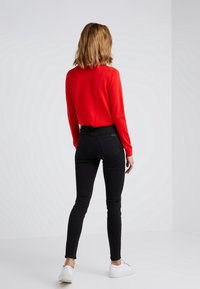Mujer con cabello hasta los hombros que lleva un suéter rojo, jeans ajustados negros y zapatillas blancas, de pie de espaldas contra un fondo liso.