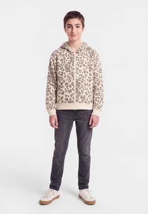 Adolescent debout, portant un sweat à capuche beige imprimé léopard avec zip, un jean gris foncé et des baskets blanches à semelles marron.