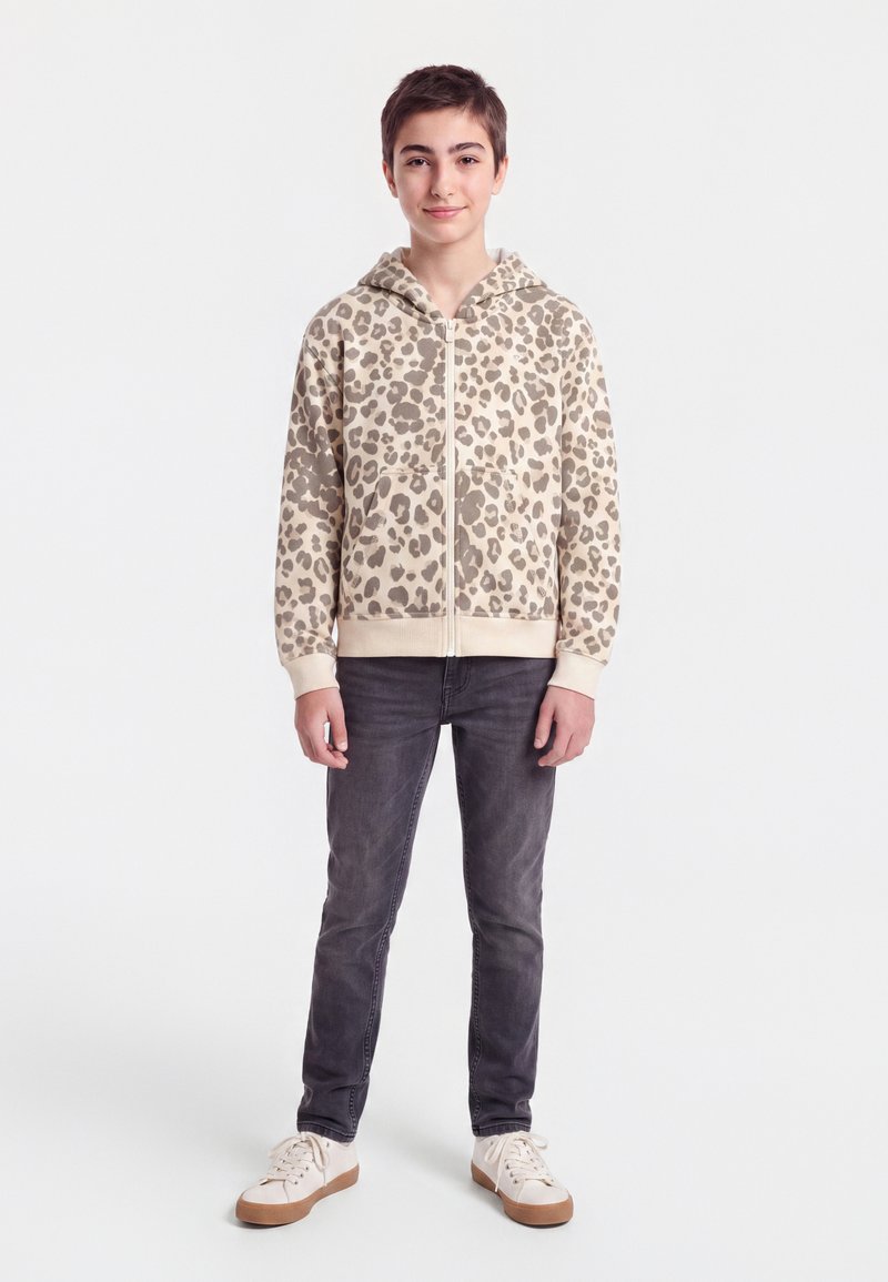 Adolescent debout, portant un sweat à capuche beige imprimé léopard avec zip, un jean gris foncé et des baskets blanches à semelles marron.