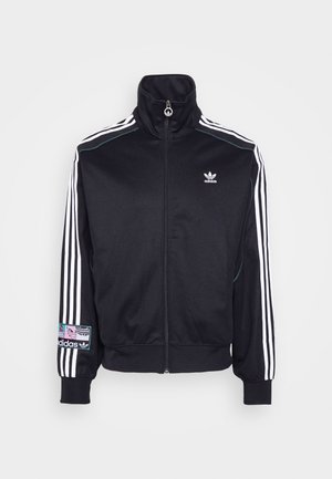 Person iført en sort Adidas trackjakke med hvide striber på ærmerne og en farverig Adidas logo-patch på venstre manchet, lynet op.