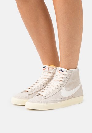 Pair of light beige suede Nike high-top sneakers worn on bare legs against a plain white background.