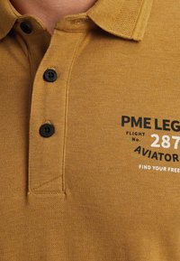 Khaki polo košile s texturovaným povrchem, se třemi černými knoflíky a potiskem značky v černé a bílé na přední straně.