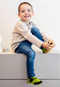 Kinderschoenen met een bovenwerk van zwart en neon groen mesh, een zachte zool en een gebogen ontwerp. Draagt door een kind dat op een grijze bank zit.