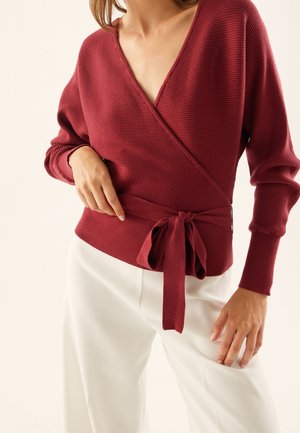 Donna che indossa un maglione bordeaux a coste, avvolto in vita e abbinato a pantaloni bianchi a vita alta, su uno sfondo semplice.