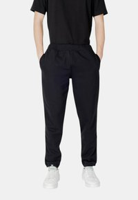 Schwarze, schmal zulaufende Jogginghosen mit elastischem Bund, seitlichen Taschen und Bündchen am Knöchel, kombiniert mit weißen Sneakers vor einem neutralen Hintergrund.