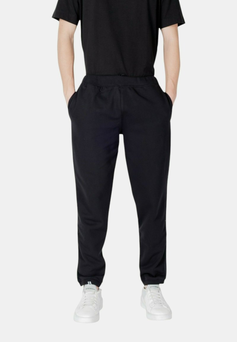 Schwarze, schmal zulaufende Jogginghosen mit elastischem Bund, seitlichen Taschen und Bündchen am Knöchel, kombiniert mit weißen Sneakers vor einem neutralen Hintergrund.