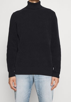 Homme portant un pull à col roulé noir à manches longues associé à un jean denim bleu clair, debout devant un fond uni.