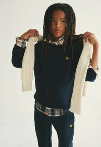 Sudadera navy con logo amarillo, superpuesta a una camisa de cuadros. El modelo sostiene un suéter de punto crema. Incluye pantalones navy con el logo a juego.