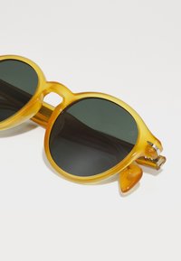 Gafas de sol redondas con un marco amarillo translúcido y lentes verdes oscuros. Incluyen un diseño de puente único y sutiles acentos texturizados en las varillas.