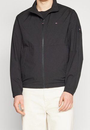 Homme portant une veste noire zippée avec un petit logo sur la poitrine et la manche, associée à un pantalon blanc cassé, debout devant un fond uni.