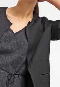 Veste noire texturée avec un col rond, arborant un design détaillé et des manches structurées, portée sur un haut sombre et scintillant.