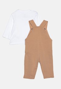 Camicia bianca a maniche lunghe abbinata a salopette color marrone chiaro con spalline regolabili e bottoni a scatto, realizzate in tessuto morbido e traspirante.
