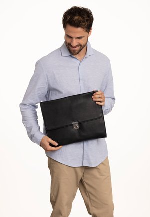 Černá kožená taška na laptop s hladkým povrchem, klopovým uzávěrem a kovovými doplňky. Držena osobou oblečenou v světle modré košili.