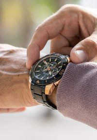 Orologio cronografo nero e oro con quadrante rotondo, dotato di un quadrante testurizzato, marcatori delle ore audaci e un bracciale in metallo, mentre viene regolato al polso.