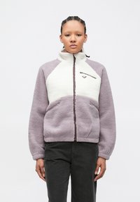 Jeune personne aux cheveux tressés portant une veste polaire lavande et blanche avec poches zippées et col montant, debout devant un fond uni.