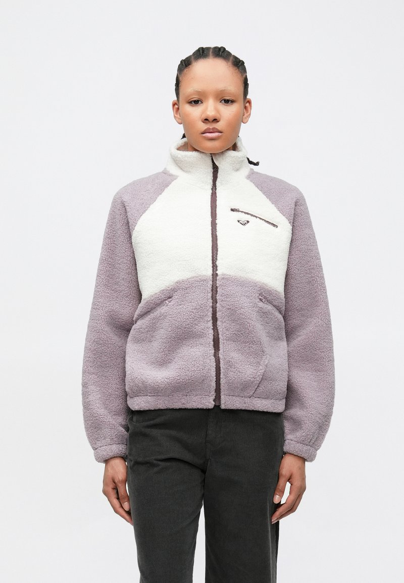 Jeune personne aux cheveux tressés portant une veste polaire lavande et blanche avec poches zippées et col montant, debout devant un fond uni.