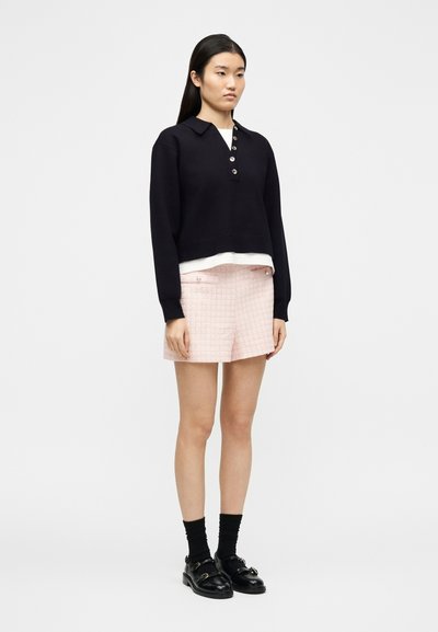 Γυναίκα που στέκεται φορώντας μαύρο μακρυμάνικο πουκάμισο με κουμπιά, λευκό εσώρουχο, ροζ υφασμάτινη βερμούδα, μαύρες κάλτσες και μαύρα μοκασίνια σε απλό φόντο.