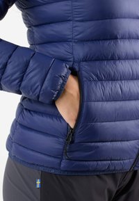 Laivastonsininen puhvitarakki, jossa on tikattu muotoilu, varustettuna vetoketjullisella sivutaskulla ja joustavilla hihansuilla. Kevyt materiaali, jossa on sileä pinta.