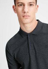 Polo shirt gris foncé en tissu texturé, avec un col, deux boutons à l'avant et un petit logo accent sur la poitrine.