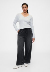 Svetlo sivi pulover z V-izrezom in dolgimi rokavi iz mehke tkanine, kombiniran s širokimi črnimi kavbojnimi hlačami z elastičnim pasom in vrvico za zavezovanje.