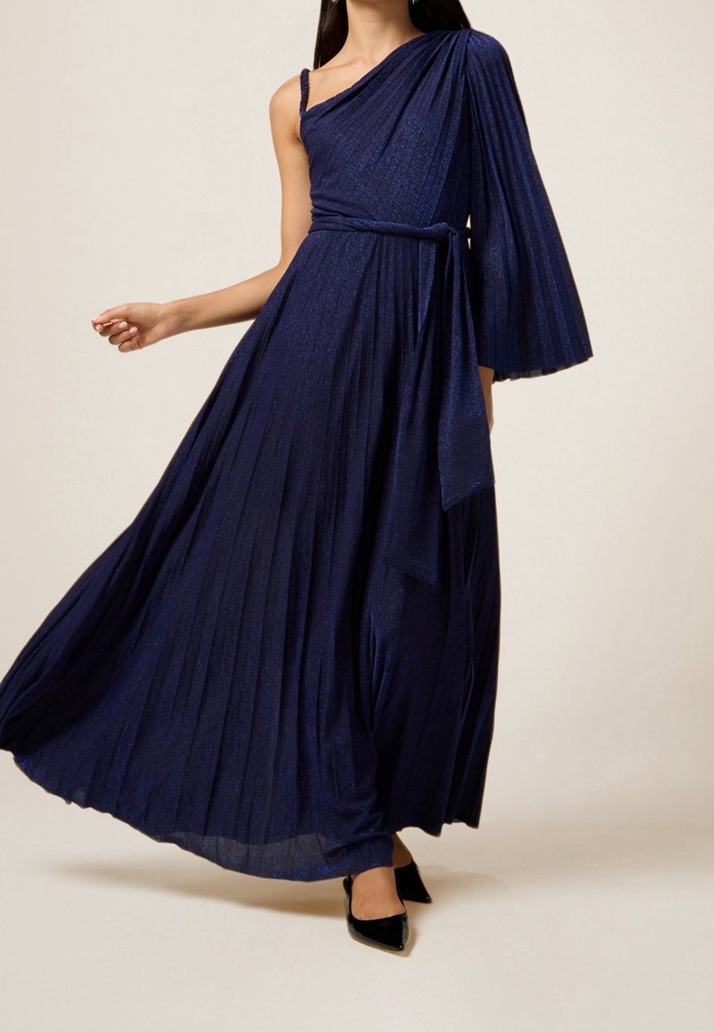 Femme portant une robe plissée asymétrique bleu foncé fluide avec une ceinture et des talons noirs pointus, debout devant un fond uni.