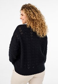 Pull en tricot noir avec un motif géométrique texturé et un ourlet côtelé. Coupe ample et manches longues, associé à un pantalon de couleur claire.