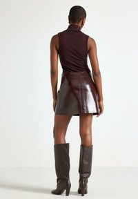 Donna che indossa un top senza maniche a collo alto, una mini gonna in pelle lucida e stivali con tacco alti fino al ginocchio, in piedi di spalle contro uno sfondo neutro.