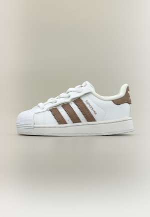 Baskets blanches Adidas Superstar avec des bandes marron, languette de talon marron et bout en gomme sur un fond neutre uni.