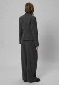 Oseba z dolgimi rjavimi lasmi, oblečena v tesno prilegajoč srajco temno sive barve in široke hlače, stoji s hrbtom obrnjenim proti sivi ozadju.