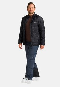 Mann in Outdoor-Bekleidung, trägt eine schwarze Daunenjacke, marineblaue Hose und weiße Skischuhe vor einem einfarbigen Hintergrund.
