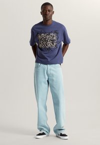 Kurzärmliges navyblaues T-Shirt mit einem kontrastierenden Grafikdesign, kombiniert mit hellblauen, locker sitzenden Jeans und schwarz-weißen Sneakern.