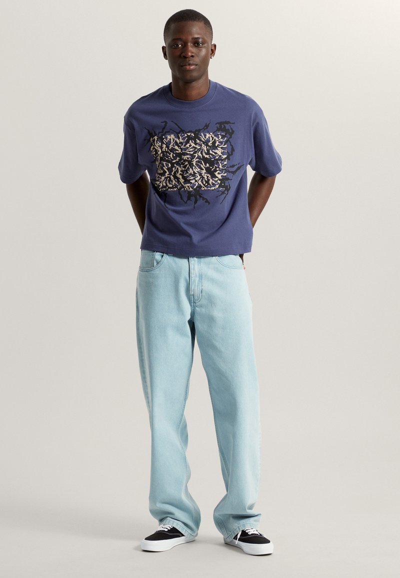 Kurzärmliges navyblaues T-Shirt mit einem kontrastierenden Grafikdesign, kombiniert mit hellblauen, locker sitzenden Jeans und schwarz-weißen Sneakern.