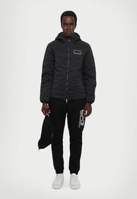 Sort quiltet jakke med hætte, der har en frontlynlås og et lille rektangulært mærke. Kombineret med sorte sweatpants og hvide sneakers.