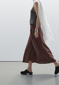 Henkilö kävelee yllään ilman hihoja harmaa ribbipaita, pitkä ruskea hame, mustat klumpit, hopeinen rannekoru ja valkoinen huivi, joka on heitetty olkapäälle.