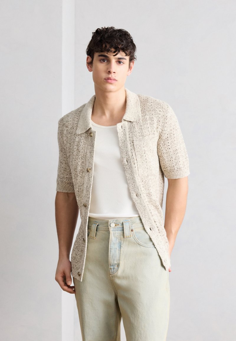 Giovane uomo con capelli ricci che indossa una maglia beige a maniche corte sopra una canottiera bianca e jeans a vita alta chiari davanti a uno sfondo semplice.