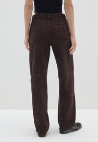 Pantaloni a gamba larga con stampa leopardata marrone e vita alta, dotati di tasche posteriori e una texture liscia. Indossati con stivali neri alla caviglia.