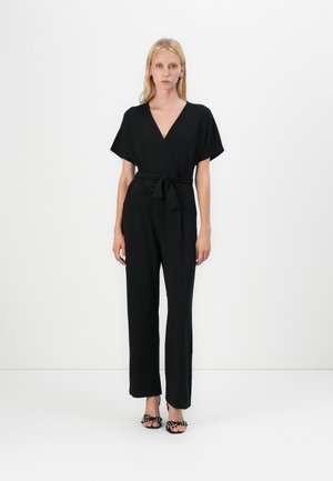 Svart jumpsuit med korte ermer, v-hals, midjebånd og rette ben, sammen med svarte sko med polka-prikker. Glatt tekstur.