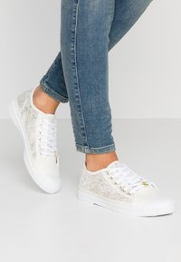Baskets blanches avec des accents argentés texturés, des lacets blancs et des semelles en caoutchouc. Présentent un design bas classique et une finition en tissu lisse.