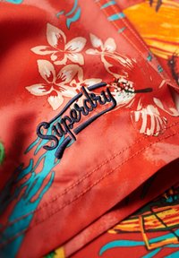 Tessuto a fantasia floreale rossa con logo "Superdry" ricamato in blu navy. Texture liscia con vivaci disegni tropicali e dettagli cuciti.