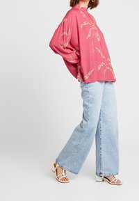 Rosa, överdimensionerad, långärmad topp med blommönster; i kombination med ljusblå, vida jeans och vita sandaler med remmar.