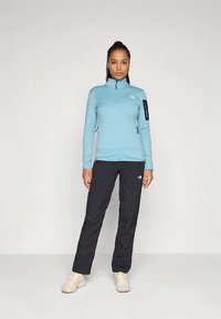 Svetlo modra jakna z zadrgo in visokim ovratnikom, črnimi poudarki in žepki z zadrgo, združena s črnimi hlačami in bež obuvali.