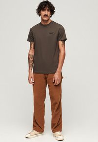Camiseta de algodón marrón con cuello redondo y logo en el pecho, combinada con pantalones de pana marrones. El modelo lleva zapatillas beige.