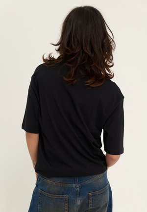 Persoon met schouderlang, donker haar, gekleed in een zwart t-shirt en blauwe spijkerbroek, met de rug naar de camera gekeerd tegen een effen lichte achtergrond.