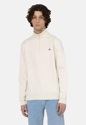 Crème zip-nekk Sweatshirt van zachte stof. Bevat een klein logo op de borst, geribbelde manchetten en een rechte zoom. Gecombineerd met lichtblauwe jeans.