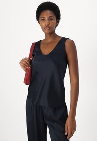 Eine Frau mit kurzen lockigen Haaren, die ein ärmelloses dunkelblaues Oberteil und eine Hose trägt, hält eine kleine rote Umhängetasche vor einem weißen Hintergrund.