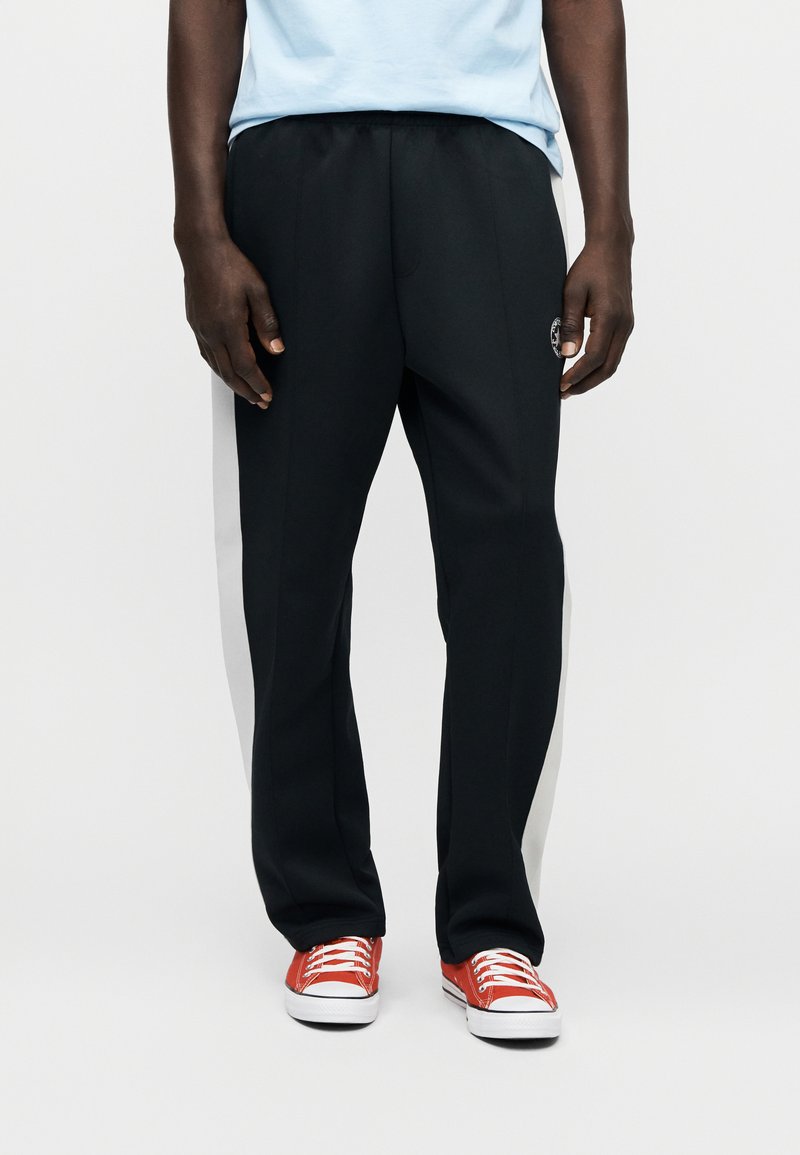 Sorte træningsbukser med hvide sidelommer, elastisk talje, diskret logo på låret, kombineret med røde sneakers.