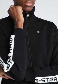 Persona vistiendo una chaqueta con cremallera negra con logotipos blancos de G-STAR en las mangas y el pecho, levantando una mano cerca de la barbilla.