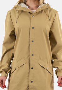 Light beige waterproof raincoat with a hood, snap buttons, flat pockets, and a textured finish. Adjustable drawstring at the neck.